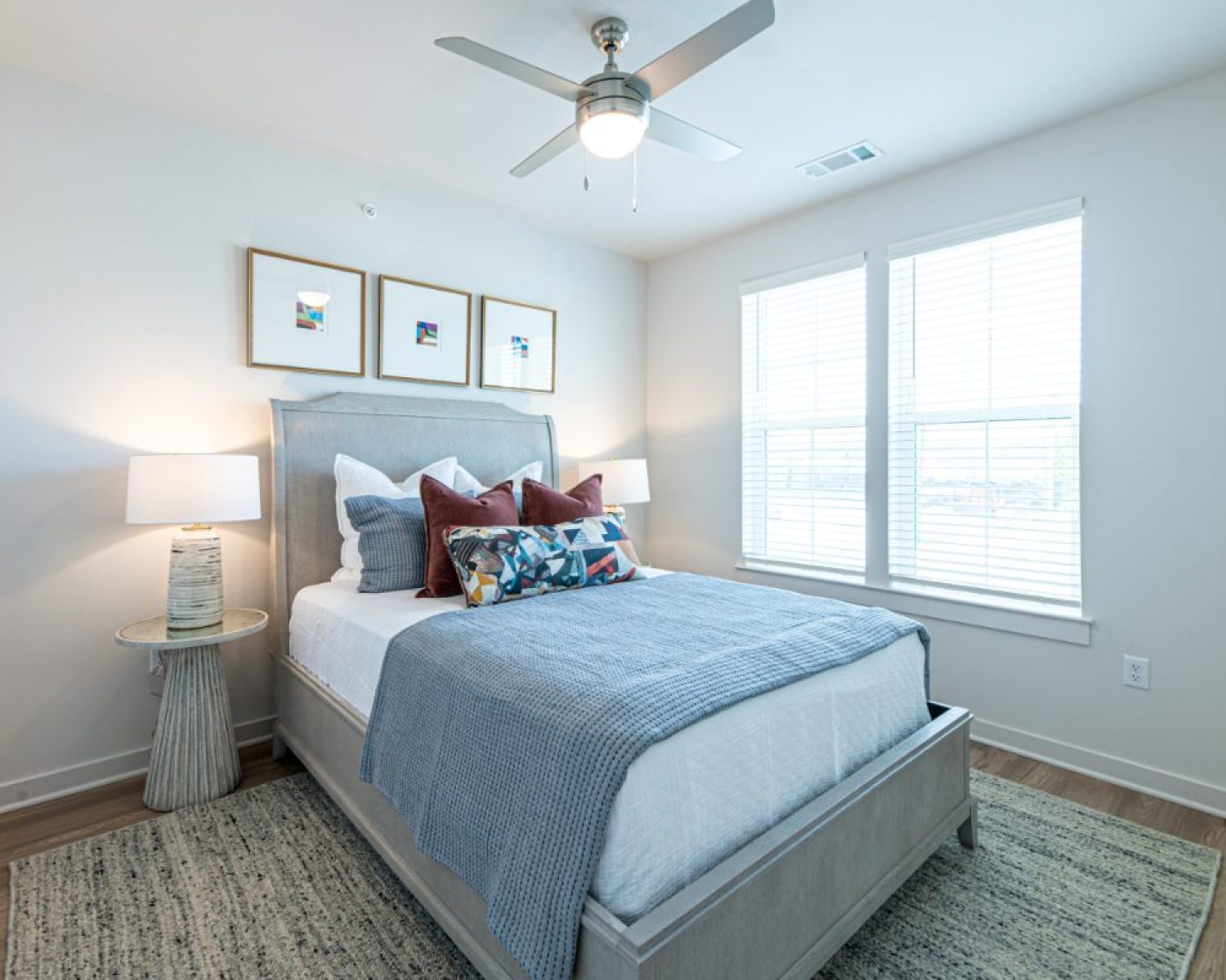 Apartments For Rent Downtown Knoxville A modern bedroom with a neatly made bed, decorative pillows, two bedside tables with lamps, framed art above the headboard, and large windows letting in natural light. Knoxville Apartments