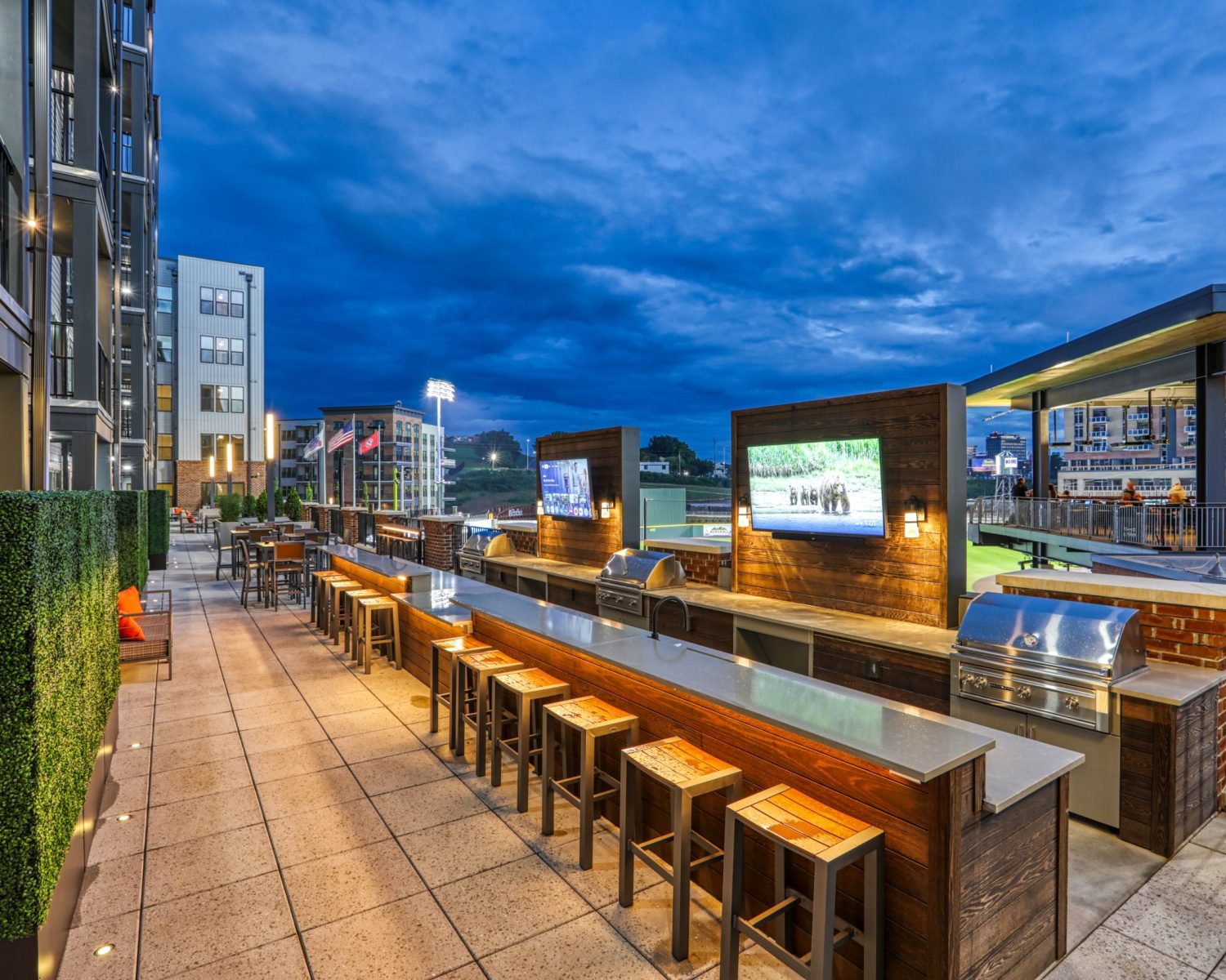 Apartments For Rent Downtown Knoxville Rooftop outdoor bar area with stools, TV screens, grills, and surrounding modern buildings at dusk. Knoxville Apartments