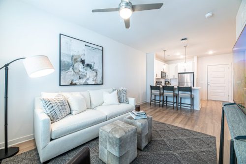 Apartments For Rent Downtown Knoxville Bright modern living room with a white sofa, abstract wall art, floor lamp, two ottomans, and an open kitchen with barstools in the background. Knoxville Apartments