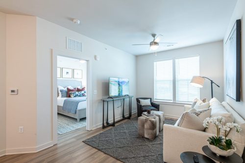 Apartments For Rent Downtown Knoxville Modern living room with a white sectional sofa, TV on a console, ottoman, floor lamp, and large window; bedroom visible through open doorway. Knoxville Apartments