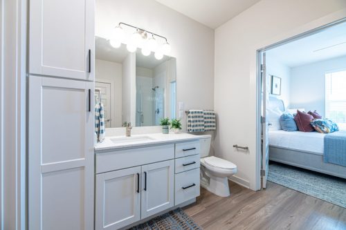 Apartments For Rent Downtown Knoxville A modern bathroom with white cabinets, a sink, a toilet, a large mirror, and a door leading to a bedroom with a neatly made bed and accent pillows. Knoxville Apartments